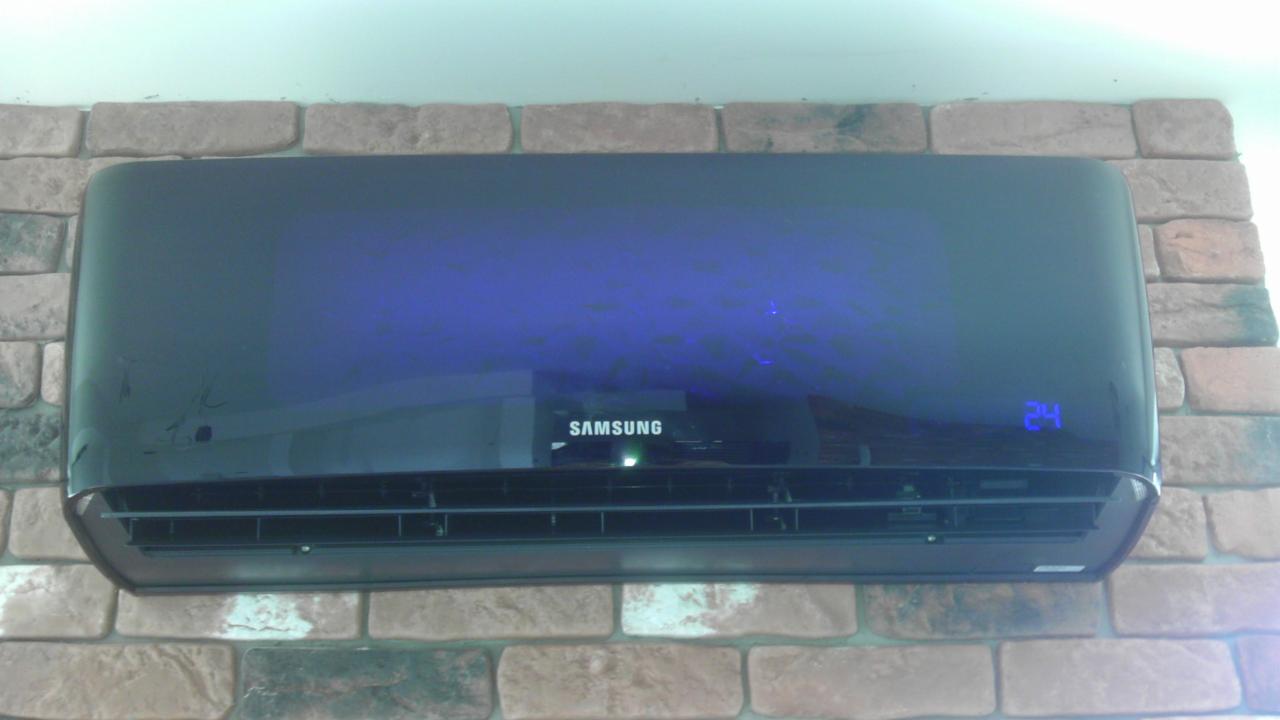 Czarny klimatyzator Samsung zamontowany na ścianie z cegły, wyświetlający temperaturę 24 stopnie Celsjusza, widoczne odbicie w błyszczącej obudowie.