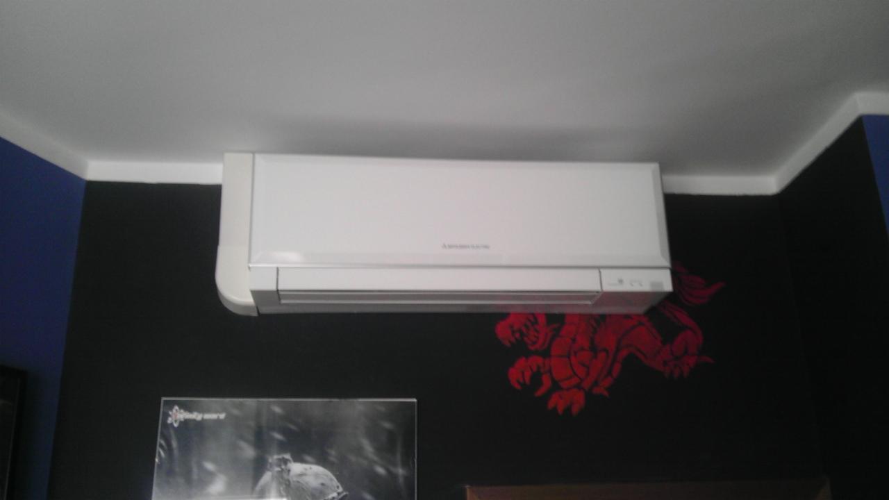Biała jednostka klimatyzacji marki Mitsubishi Electric zamontowana na czarnej ścianie z czerwonym motywem smoka, widoczny fragment sufitu i niebieskiej ściany w rogu.
