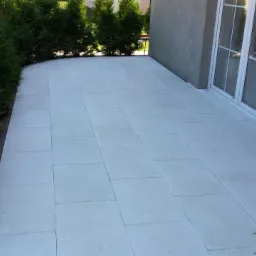 Jasnoszara kostka brukowa ułożona na tarasie przy budynku, z widocznymi krzewami po bokach i tujami w tle. Widok z perspektywy lekko z góry.