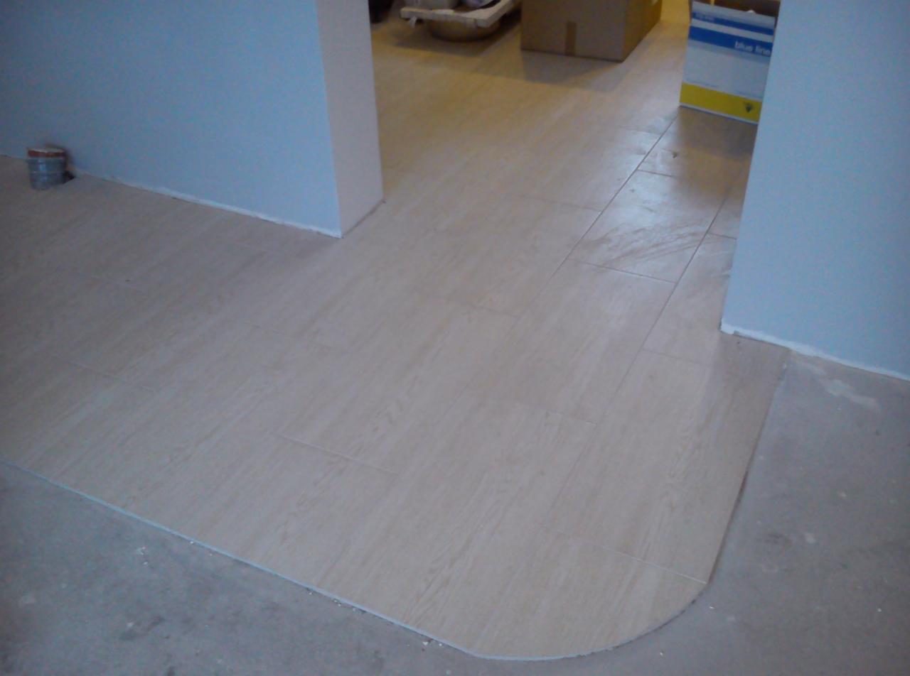 Ułożona jasna terakota w przejściu między pomieszczeniami, z zaokrąglonym narożnikiem na styku z surową posadzką.