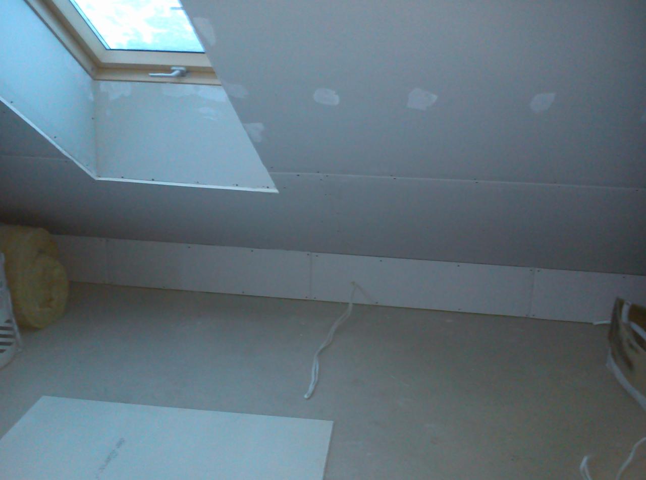 Poddasze w trakcie zabudowy płytami gipsowo-kartonowymi, widoczne łączenia płyt na suficie i ścianach, okno dachowe u góry kadru, fragment wełny mineralnej w rogu.