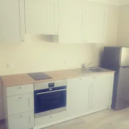 Jasna, nowa kuchnia z białymi szafkami, drewnianym blatem, płytą indukcyjną i srebrną lodówką w rogu.