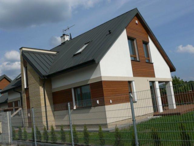 Nowoczesny dom jednorodzinny z elewacją z desek drewnianych w kolorze jasnego brązu, kontrastującą z białym tynkiem i grafitowym dachem, widziany zza metalowego ogrodzenia.