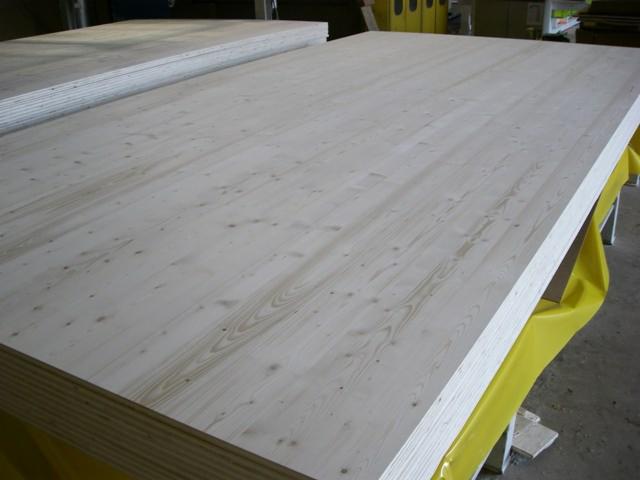 Stos płyt warstwowych z jasnego drewna iglastego, widoczne krawędzie i usłojenie, zabezpieczone folią podczas transportu.