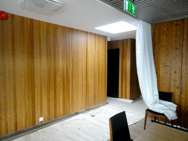 Wnętrze wyłożone pionowymi panelami z jasnego drewna z widocznym zabezpieczeniem podłogi folią malarską, krzesło z narzuconą białą tkaniną oraz znak ewakuacyjny pod sufitem.
