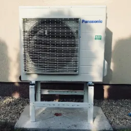 Biała jednostka zewnętrzna pompy ciepła Panasonic na metalowym stelażu, umieszczona na betonowej podstawie obok elewacji budynku, z widocznym logo i oznaczeniem czynnika chłodniczego R32.