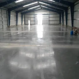 Błyszcząca, szara posadzka przemysłowa w hali magazynowej z widocznym sprzętem do polerowania i niebieskim pojemnikiem. Metalowa konstrukcja dachu i ścian.