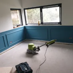 Pokój w trakcie remontu z pomalowanymi na niebiesko panelami ściennymi, maszyna do polerowania podłóg stoi na jasnej posadzce, widoczny czarny kufer z narzędziami i butelka na parapecie okna.