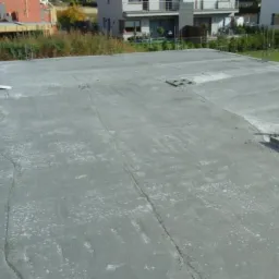 Wylany betonowy strop budynku w trakcie budowy, widoczne zbrojenia, szalunki i ślady po narzędziach, w tle zabudowa jednorodzinna.