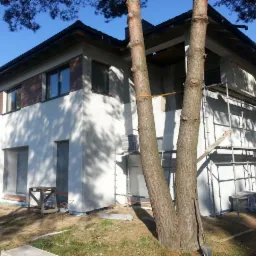 Surowy stan domu murowanego z częściowo wykonaną elewacją drewnopodobną na piętrze, widoczne rusztowania i przygotowane materiały budowlane wokół, ujęcie z dołu przez dwa drzewa.