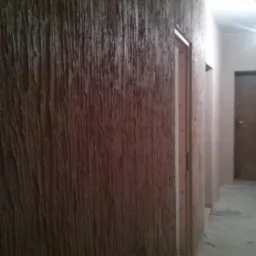 Brązowa ściana z pionowymi, teksturowanymi smugami przypominającymi korę drzewa, widok na korytarz z drzwiami w tle.