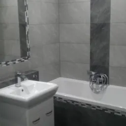 Wykończona łazienka w odcieniach szarości z umywalką na szafce, wanną obudowaną płytkami i lustrem z mozaikową ramą.