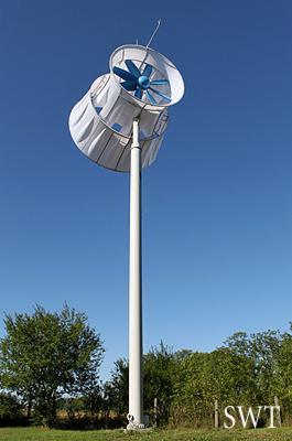 Dyfuzorowa turbina wiatrowa SWT-15 z niebieskimi łopatami, zamontowana na wysokim, białym słupie na tle błękitnego nieba i zieleni drzew.