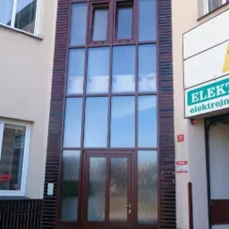 Elewacja budynku z wysokimi, brązowymi oknami i drzwiami wejściowymi, obramowanymi ciemną cegłą klinkierową, z widocznym szyldem firmy obok.