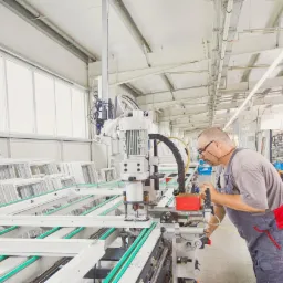 Pracownik obsługuje maszynę do produkcji ram okiennych z białego profilu, widoczne ramy w kolejce do obróbki i okna w tle.