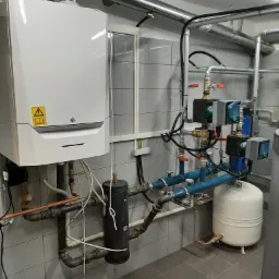 Biały piec gazowy De Dietrich w kotłowni w Rybniku, z widocznymi rurami miedzianymi, czarnym filtrem i białym zbiornikiem wyrównawczym, na tle szarych płytek.