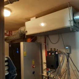 Wnętrze kotłowni z zainstalowanym nowoczesnym piecem, zbiornikiem buforowym i systemem wentylacji z szarymi rurami, widoczne liczne przewody, zawory i osprzęt hydrauliczny oraz umywalka.