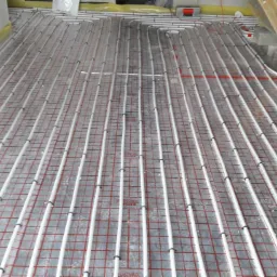 Białe rury ogrzewania podłogowego ułożone równolegle na folii z czerwoną siatką, widok z góry, surowy stan wykończenia podłogi.