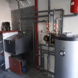 Nowoczesna kotłownia z zasobnikiem ciepłej wody, kotłem na paliwo stałe z podajnikiem i instalacją hydrauliczną z rur miedzianych i stalowych, widoczne pompy obiegowe i manometry.