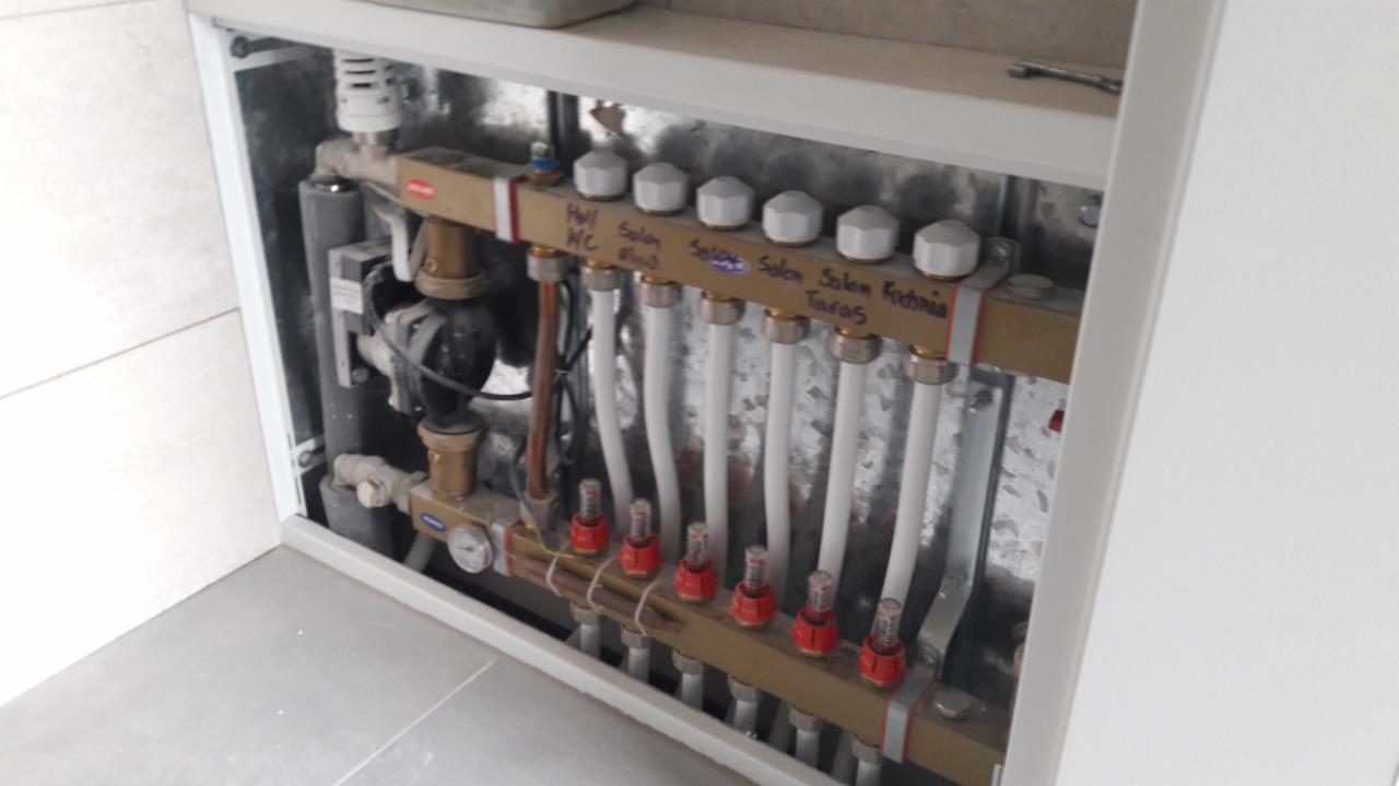 Rozdzielacz ogrzewania podłogowego w zabudowie, widoczne białe rury doprowadzające wodę do poszczególnych pomieszczeń: Hall WC, Salon, Sypialnia, Kuchnia, z opisami pomieszczeń naniesionymi markerem.