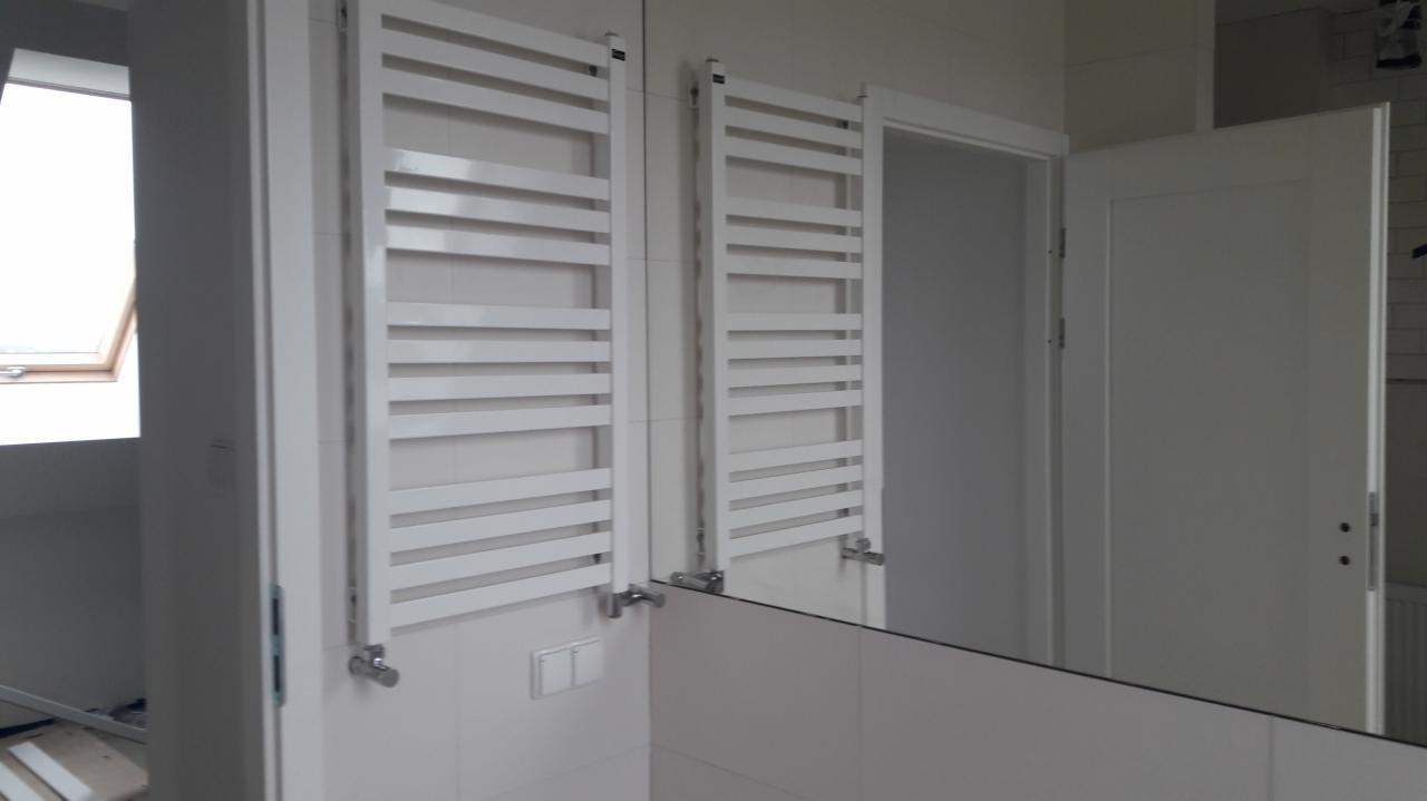 Dwa białe, drabinkowe grzejniki łazienkowe zamontowane na ścianie z białych płytek, widoczne odbicie w lustrze, białe drzwi i okno dachowe w tle.