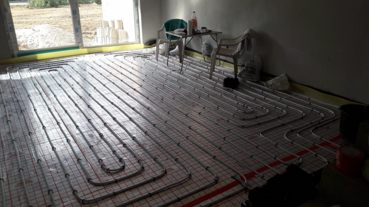 Rozłożona instalacja ogrzewania podłogowego z rurkami na folii z siatką w pomieszczeniu w trakcie remontu, widoczne narzędzia pracy i prowizoryczne meble.