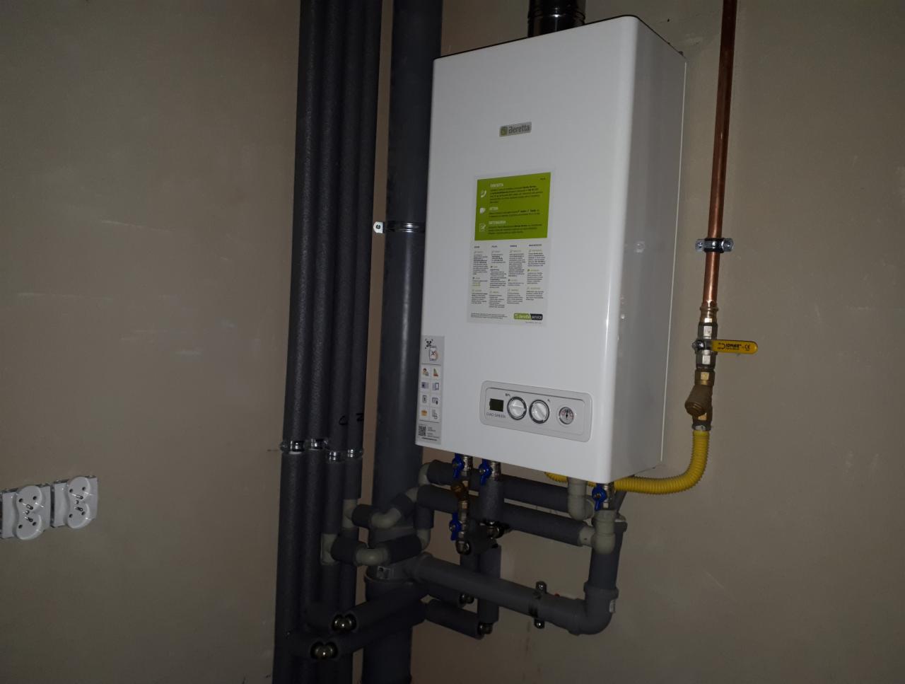 Biały, wiszący kocioł gazowy Beretta z żółtym przewodem gazowym i szarymi rurami instalacji CO, zamontowany na beżowej ścianie. Widoczne pionowe rury w izolacji oraz gniazdka elektryczne na ścianie.