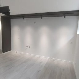 Daniel Grela - Nowoczesne wnętrze z jasną podłogą, białymi ścianami i oknem. Widoczne elementy oświetlenia sufitowego i instalacje. Minimalistyczny design.