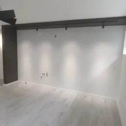 Nowoczesne wnętrze z jasną podłogą, białymi ścianami i oknem. Widoczne elementy oświetlenia sufitowego i instalacje. Minimalistyczny design.