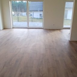 Daniel Grela - Jasne, przestronne wnętrze z drewnianą podłogą i dużymi oknami, widok na ogród. Dwa panele sterowania na ścianie. Stan wykończenia.