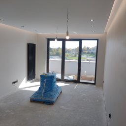 Daniel Grela - Wnętrze w trakcie wykańczania: gładkie, białe ściany, grzejnik, okno balkonowe. Podłoga przygotowana pod wylewkę, zabezpieczone materiały budowlane.