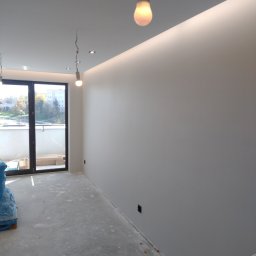 Daniel Grela - Wnętrze po malowaniu: gładkie, jasne ściany, widoczne niedoskonałości podłogi i surowe żarówki. Balkon z widokiem na zieleń w tle.