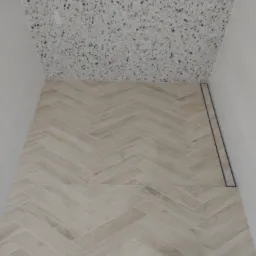 Wykończona łazienka z kremowymi płytkami ułożonymi w jodełkę na podłodze i ścianach, oraz mozaiką na suficie. Widoczny odpływ liniowy.