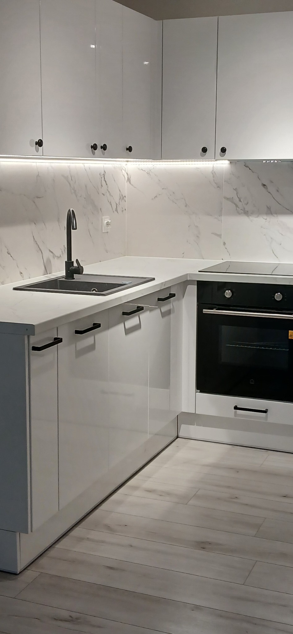 Nowoczesna, biała kuchnia z czarną armaturą i marmurowym backsplashiem podświetlonym LED, prezentująca minimalistyczny design i funkcjonalność.