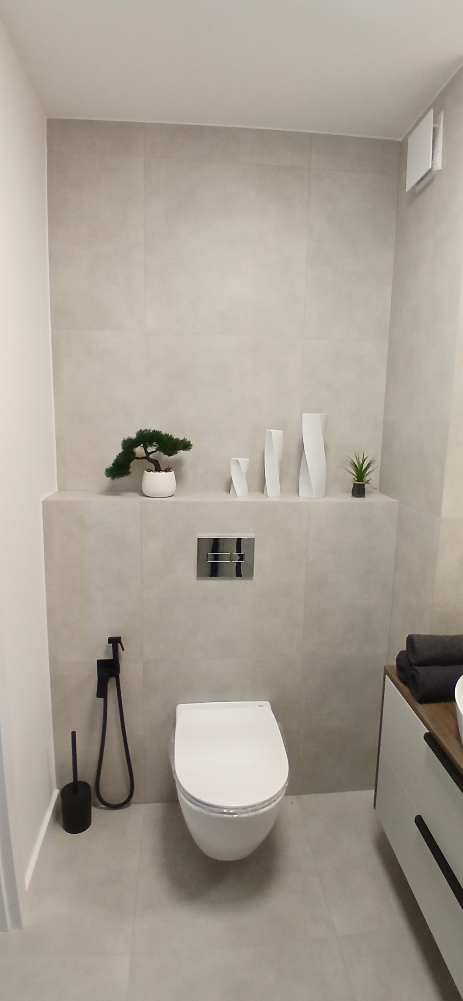 Nowoczesna toaleta w jasnych płytkach, z ozdobnymi roślinami i minimalistyczną armaturą, tworząca spójną i elegancką przestrzeń łazienkową.