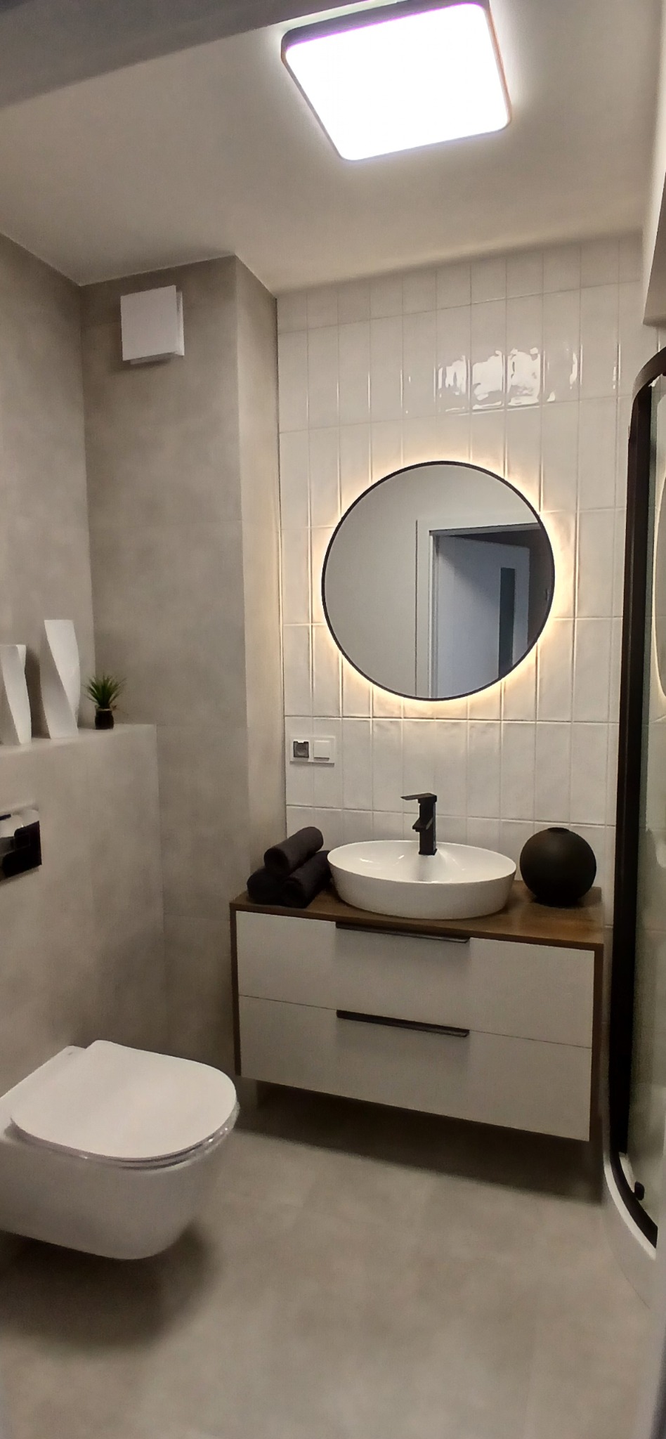 Nowoczesna łazienka z okrągłym lustrem podświetlanym LED, białą umywalką nablatową i wiszącą toaletą. Minimalistyczny design w szaro-białej kolorystyce.