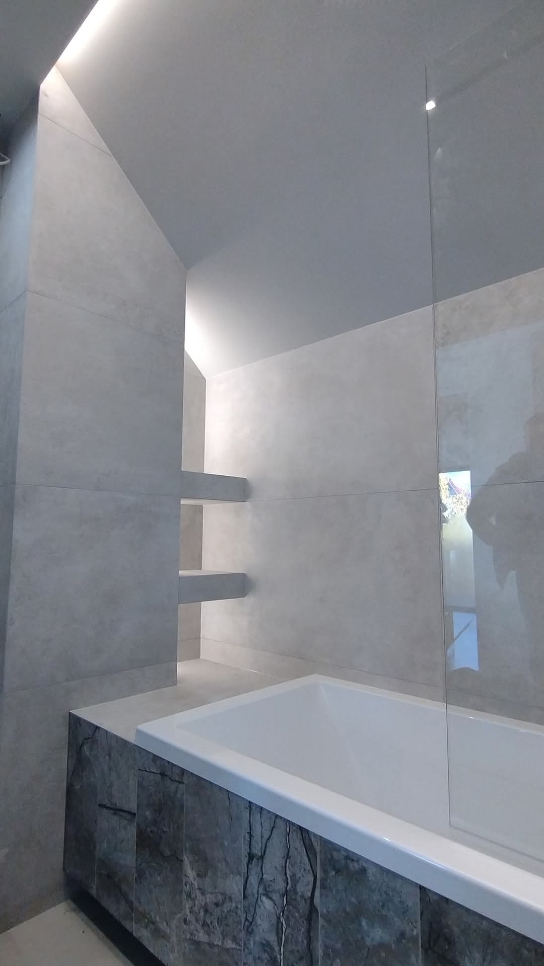 Nowoczesna łazienka z wanną obudowaną kamieniem, szklaną ścianką i półkami wnękowymi, podświetlona ledami. Minimalistyczny design, jasne płytki.