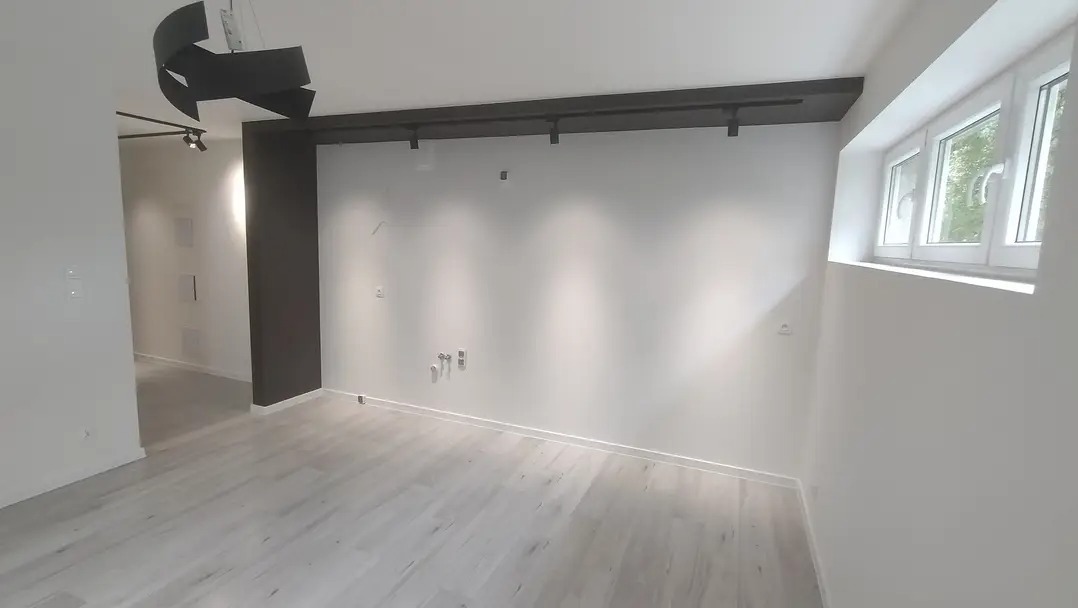 Nowoczesne wnętrze z jasną podłogą, białymi ścianami i oknem. Widoczne elementy oświetlenia sufitowego i instalacje. Minimalistyczny design.