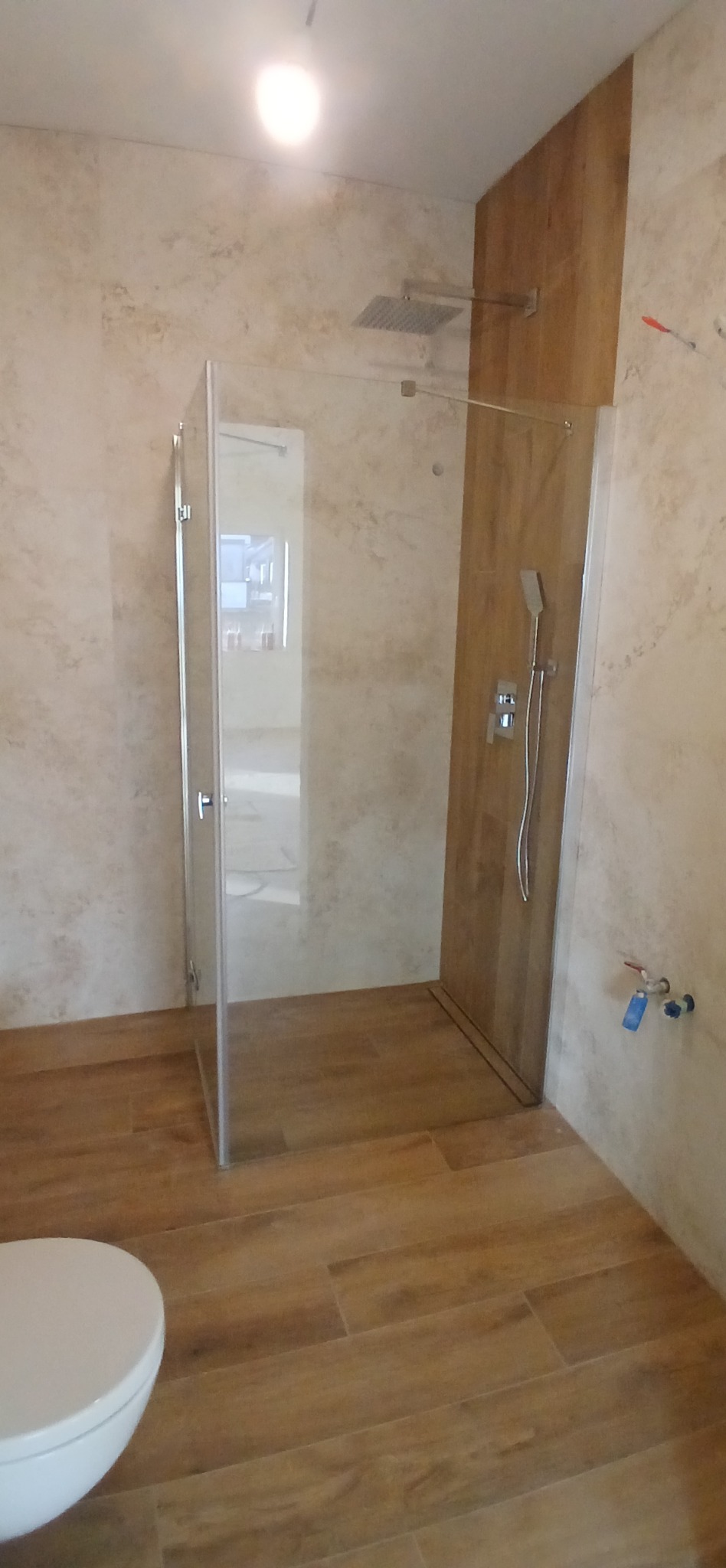 Wykończona łazienka z kabiną typu walk-in, wykończona w beżowych odcieniach z drewnopodobną podłogą i częściowo drewnianą ścianą. Widoczna armatura i toaleta.