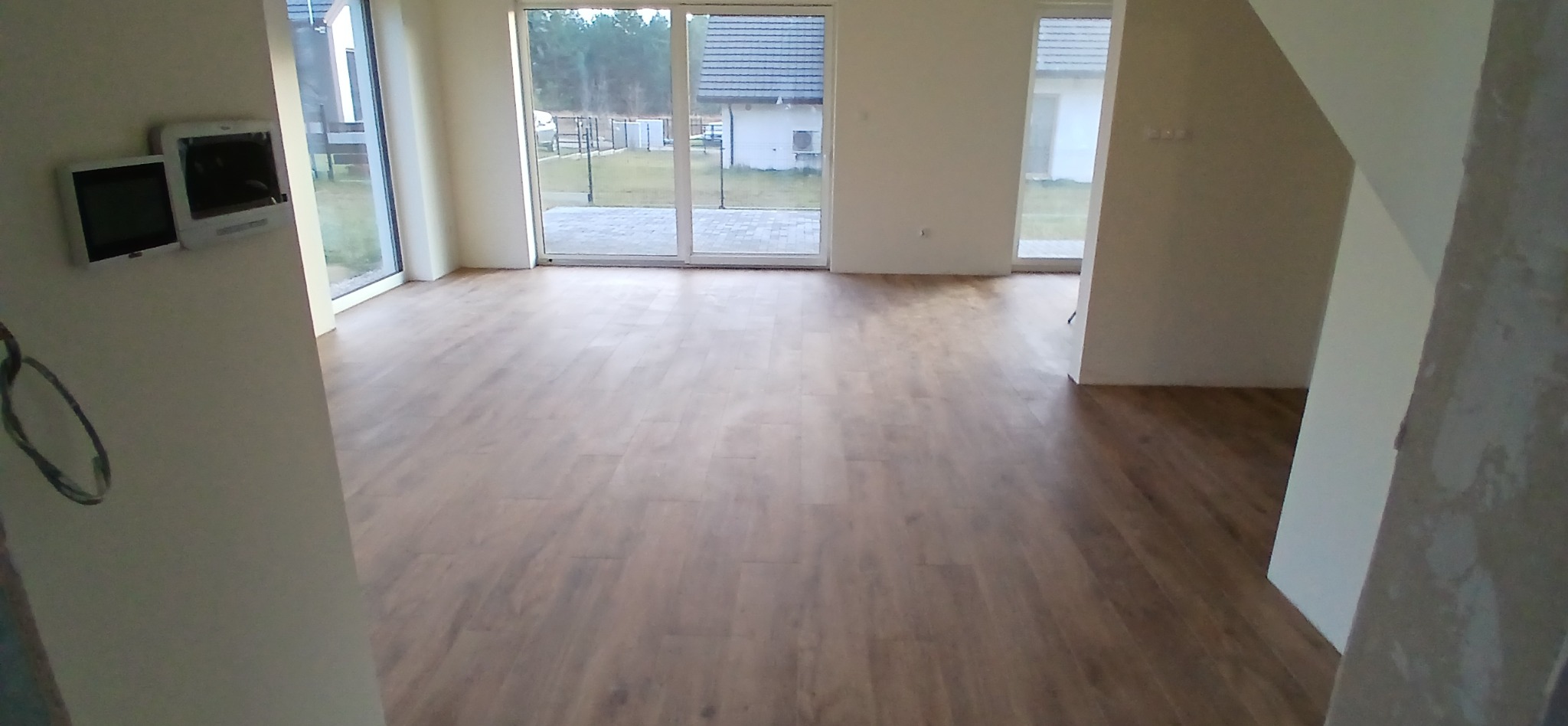 Jasne, przestronne wnętrze z drewnianą podłogą i dużymi oknami, widok na ogród. Dwa panele sterowania na ścianie. Stan wykończenia.