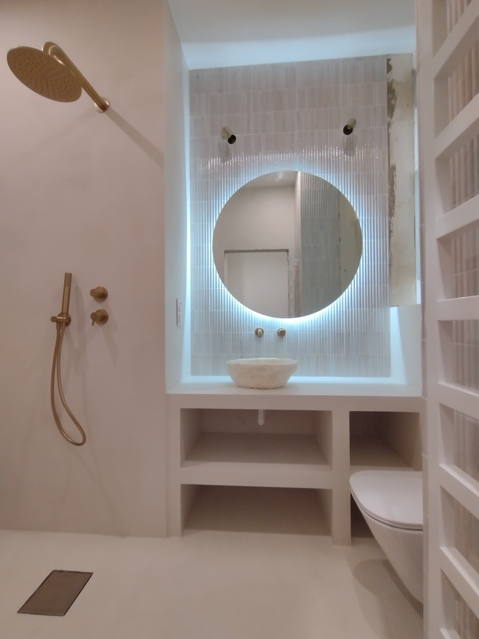 Minimalistyczna łazienka w bieli z okrągłym lustrem podświetlanym LED, armaturą w kolorze złota i płytkami z delikatnym wzorem. Nowoczesny design z elementami naturalnego kamienia.