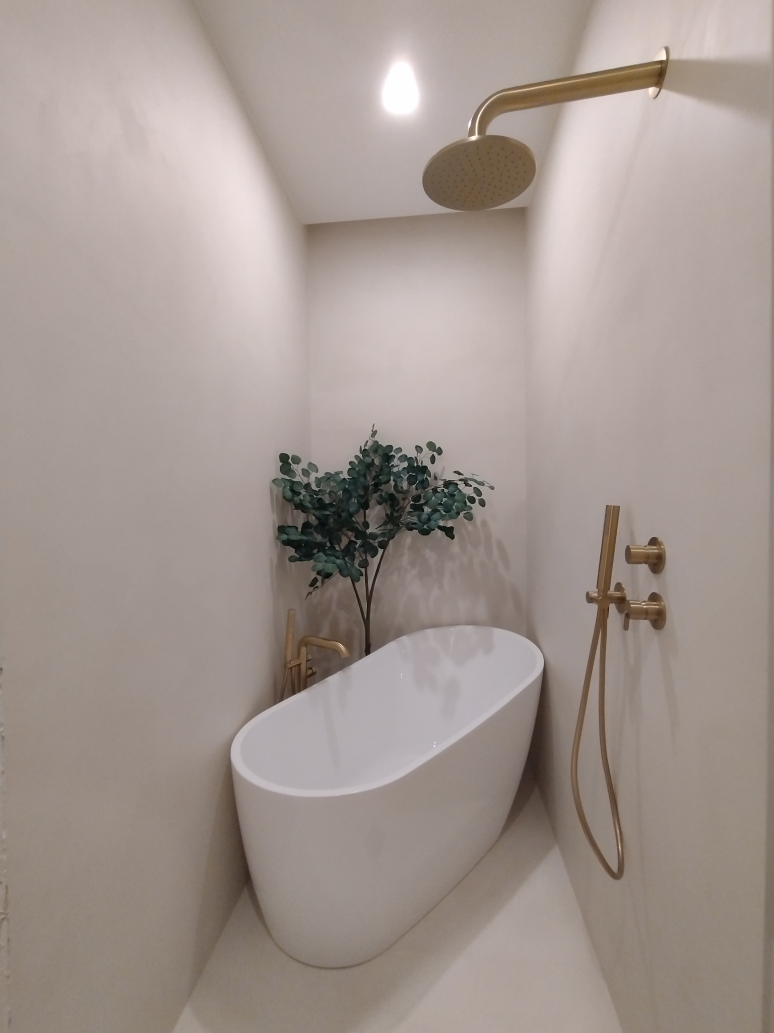 Minimalistyczna łazienka z wolnostojącą wanną, złotą armaturą i rośliną doniczkową, oświetlona punktowo, ukazująca elegancję wykończenia wnętrza.