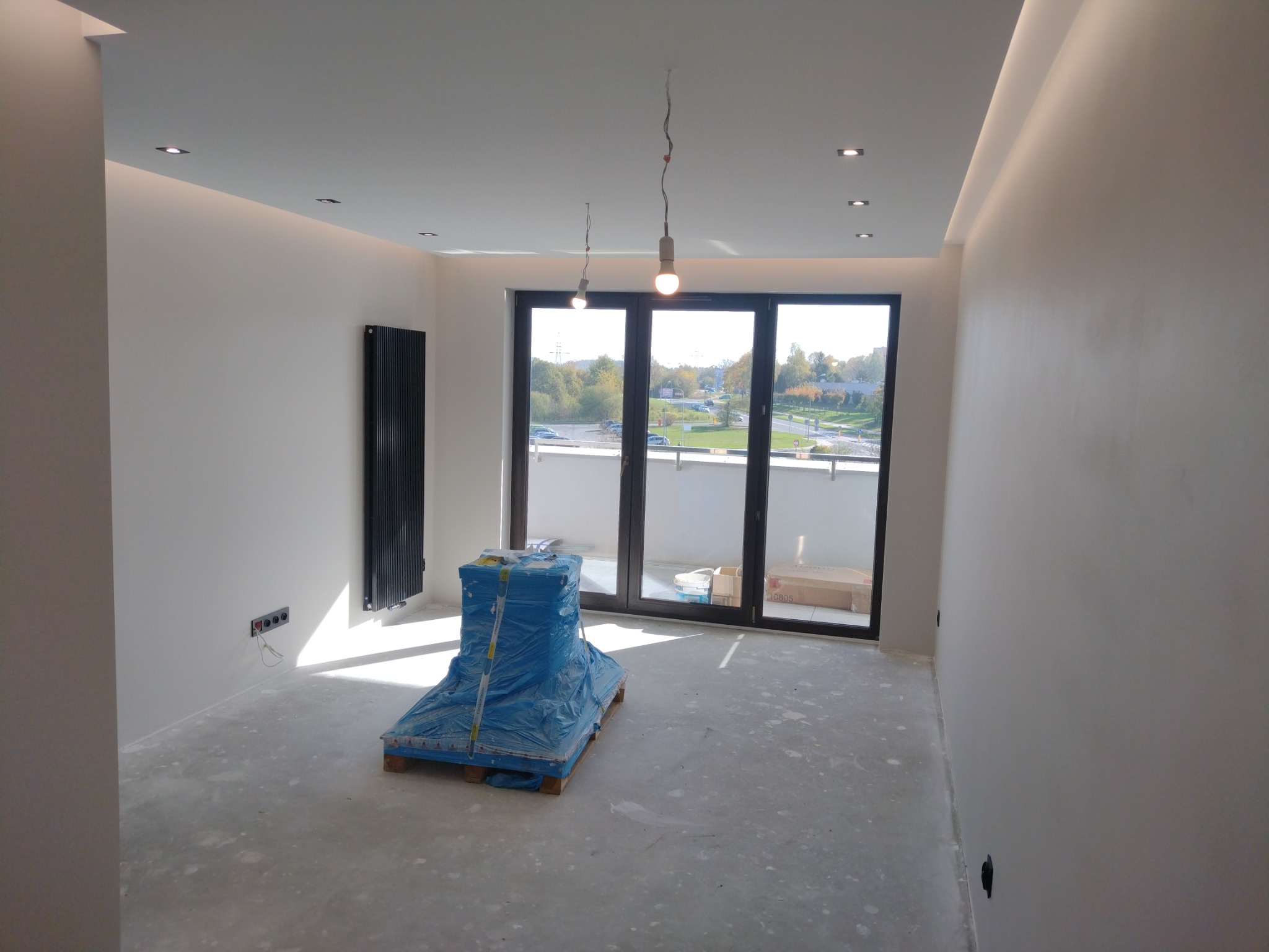 Wnętrze w trakcie wykańczania: gładkie, białe ściany, grzejnik, okno balkonowe. Podłoga przygotowana pod wylewkę, zabezpieczone materiały budowlane.