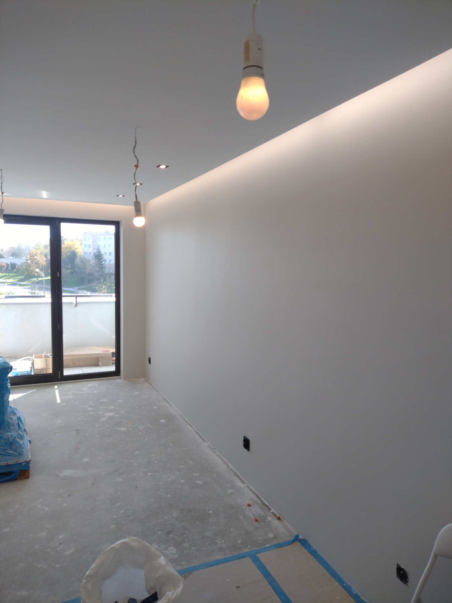 Wnętrze po malowaniu: gładkie, jasne ściany, widoczne niedoskonałości podłogi i surowe żarówki. Balkon z widokiem na zieleń w tle.