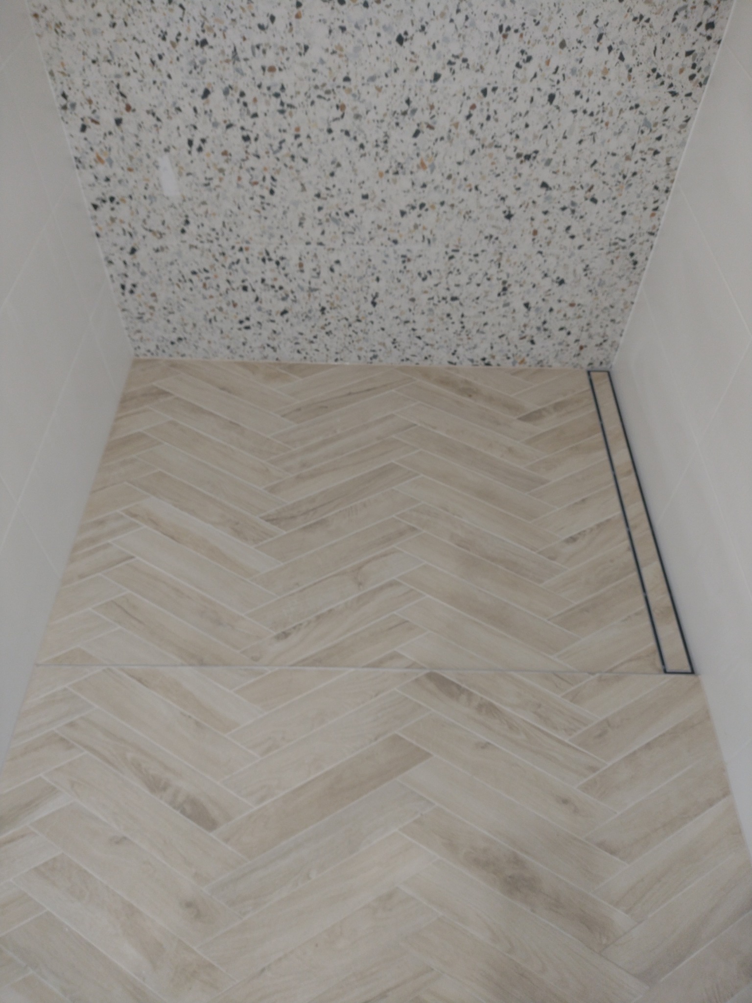 Wykończona łazienka z kremowymi płytkami ułożonymi w jodełkę na podłodze i ścianach, oraz mozaiką na suficie. Widoczny odpływ liniowy.