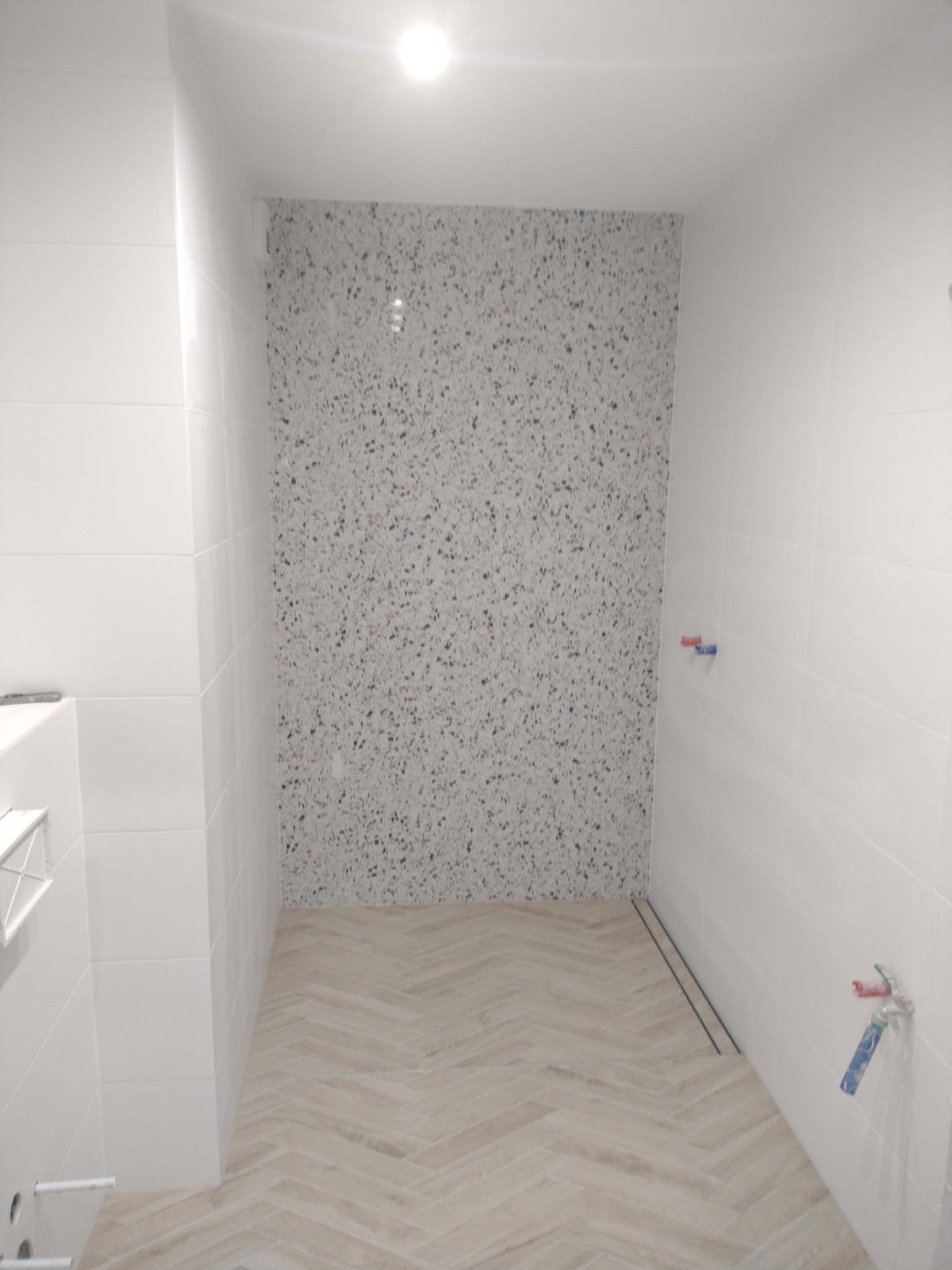 Wykończona łazienka z białymi płytkami i podłogą w jodełkę. Ściana prysznicowa z mozaiką w odcieniach szarości. Widoczne elementy instalacji wodnej.