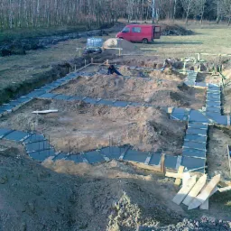 Wykop pod fundamenty z betonowymi ławami, widoczne szalunki i wystające pręty zbrojeniowe. W tle czerwony bus i zbiornik na wodę.