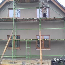 Prace tynkarskie na elewacji domu jednorodzinnego z użyciem rusztowania. Widoczny pracownik na rusztowaniu z liną. Tynk w kolorze szarym.