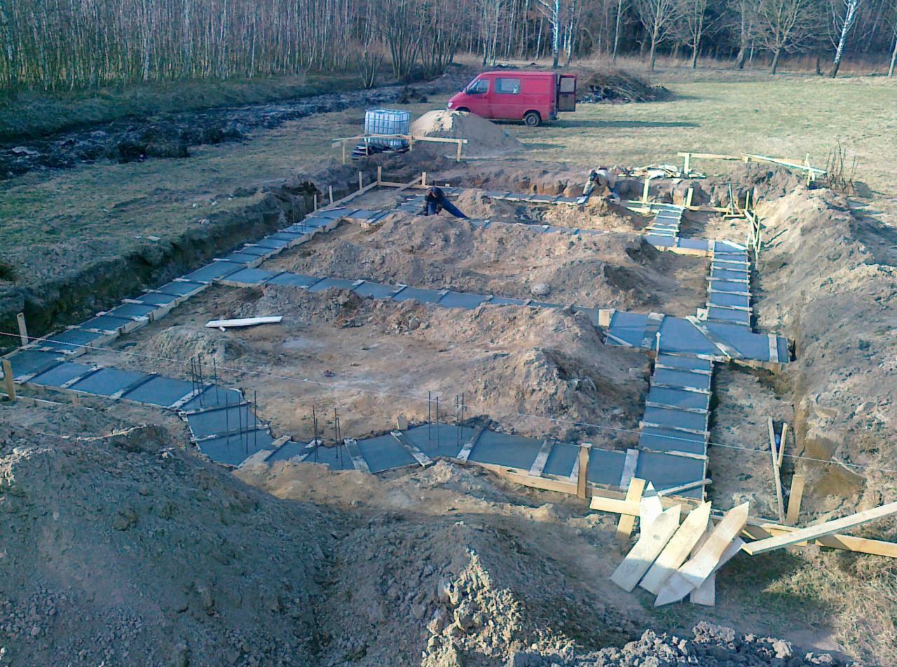 Wykop pod fundamenty z betonowymi ławami, widoczne szalunki i wystające pręty zbrojeniowe. W tle czerwony bus i zbiornik na wodę.