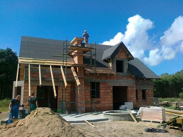 Budowa domu z czerwonej cegły: murarz na rusztowaniu przy kominie, widoczne elementy dachu, okna, drewniane belki i zaplecze budowy z dwoma pracownikami.